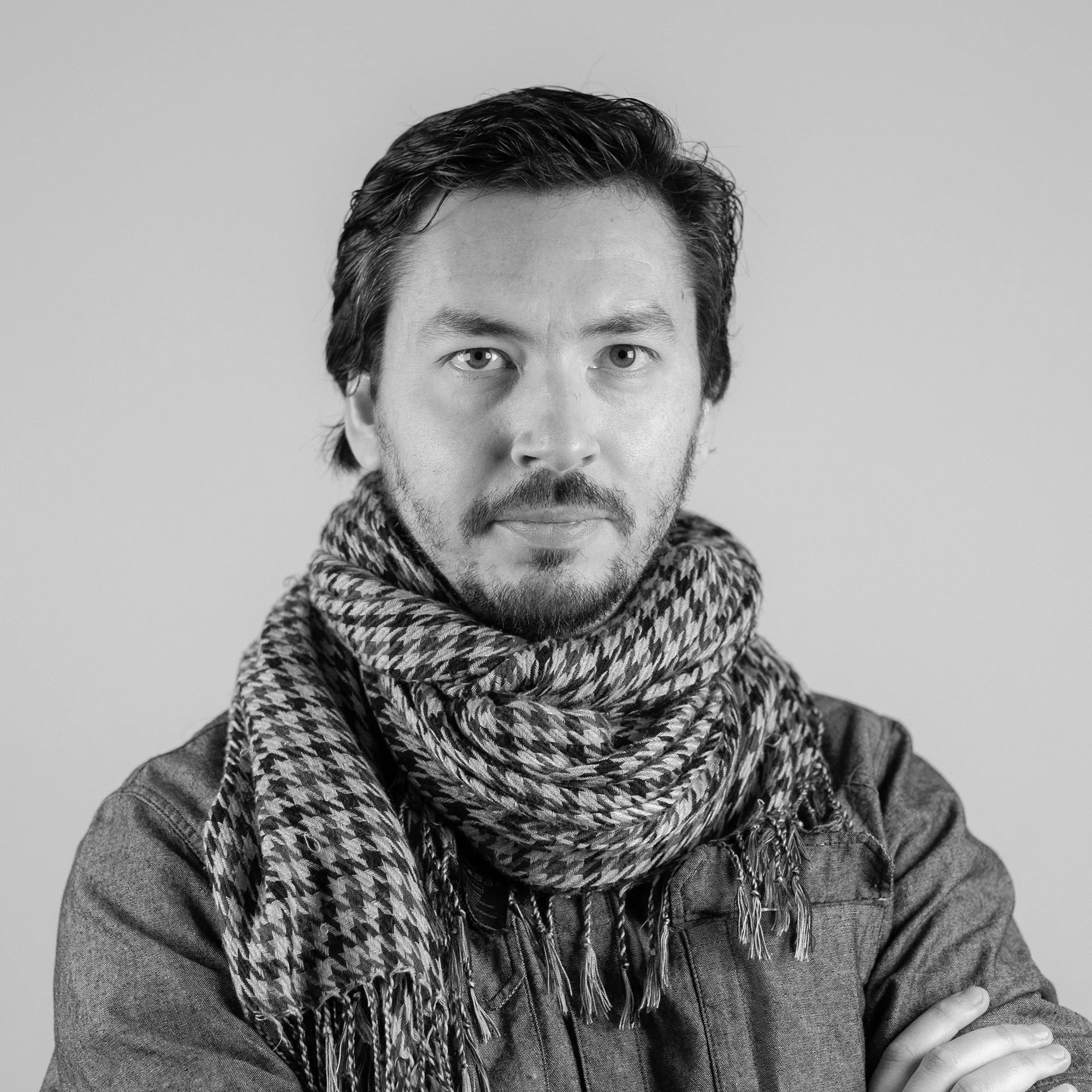 Retrato de un hombre con barba y cabello oscuro, usando una bufanda a cuadros y una chaqueta, en blanco y negro.
