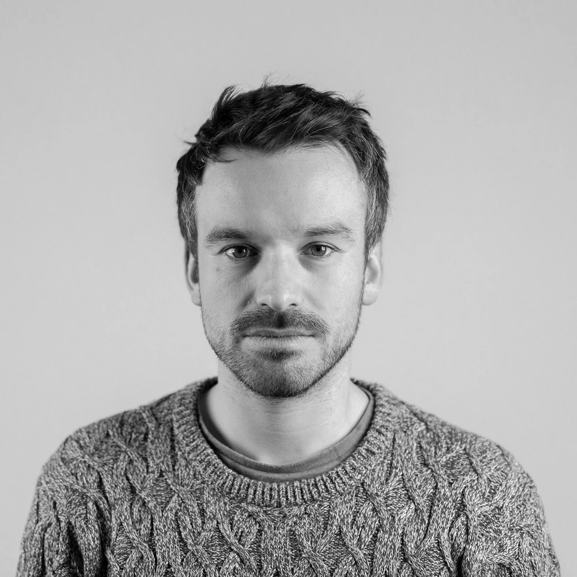 Retrato en blanco y negro de un hombre con barba y cabello cortado, usando un suéter, mirando a la cámara con expresión neutral.