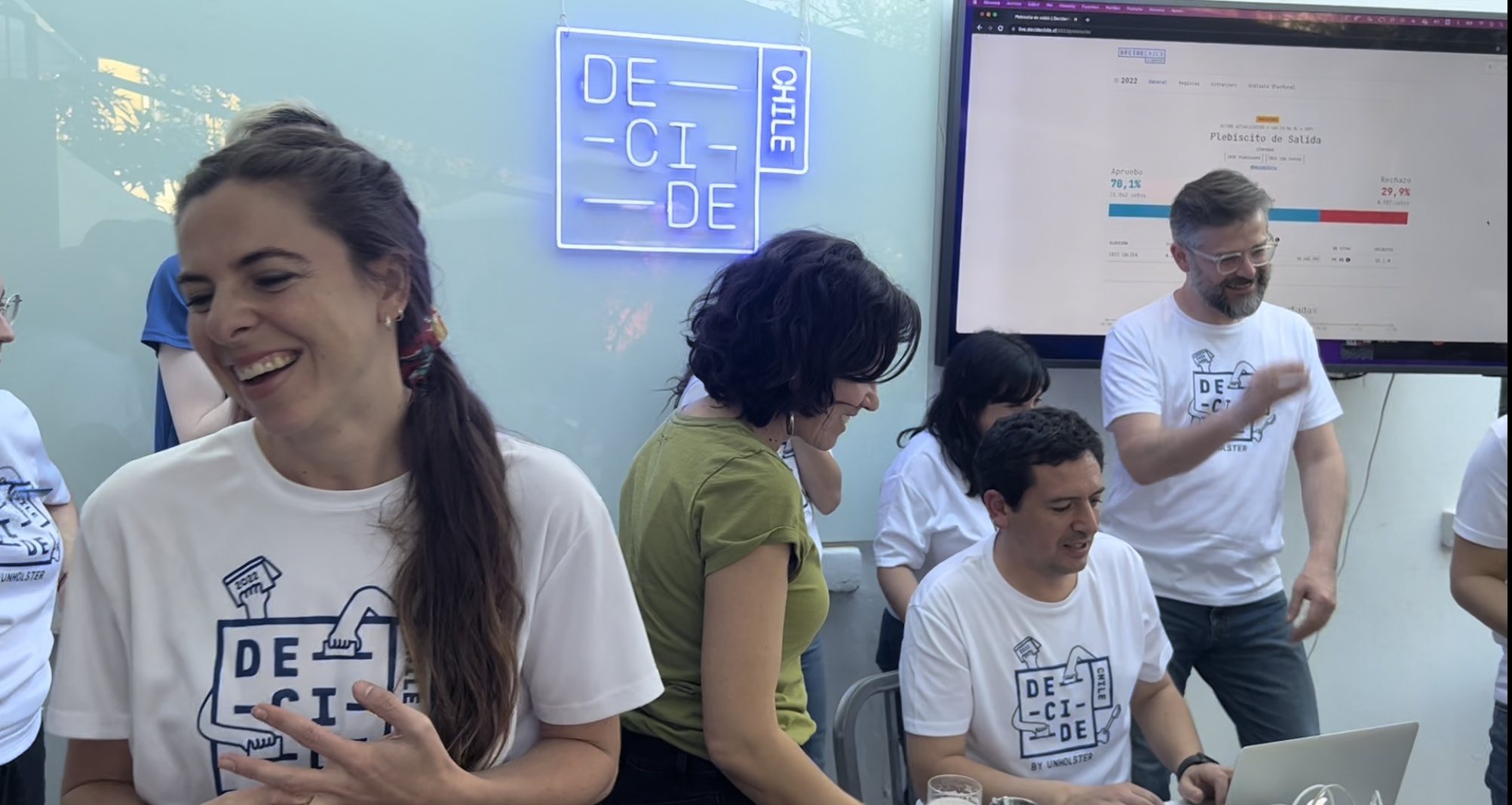 El equipo con poleras Decide Chile frente al letrero de neón con el logotipo y una pantalla con los resultados del plebiscito de salida.